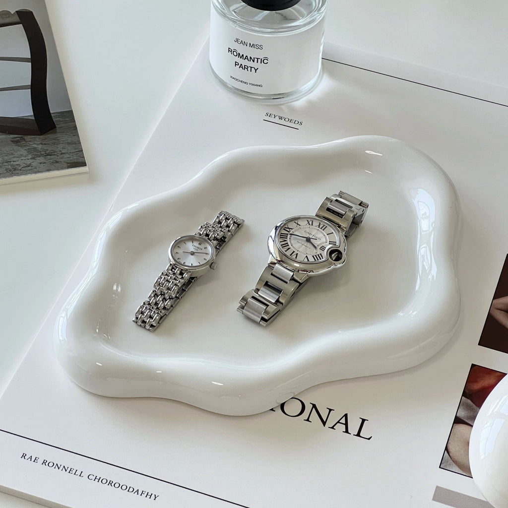 Two silver-toned stainless steel wristwatches with white dials and roman numerals on a ceramic cloud tray