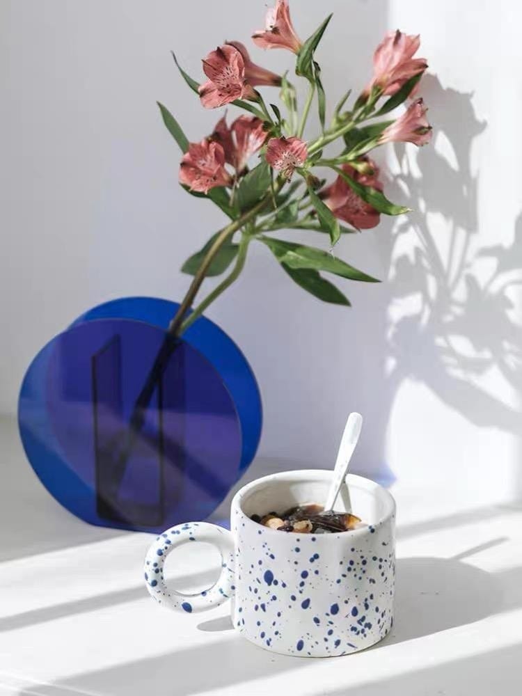 White ceramic dumbo mug with blue speckles and circular handle filled with dark liquid and holding a spoon