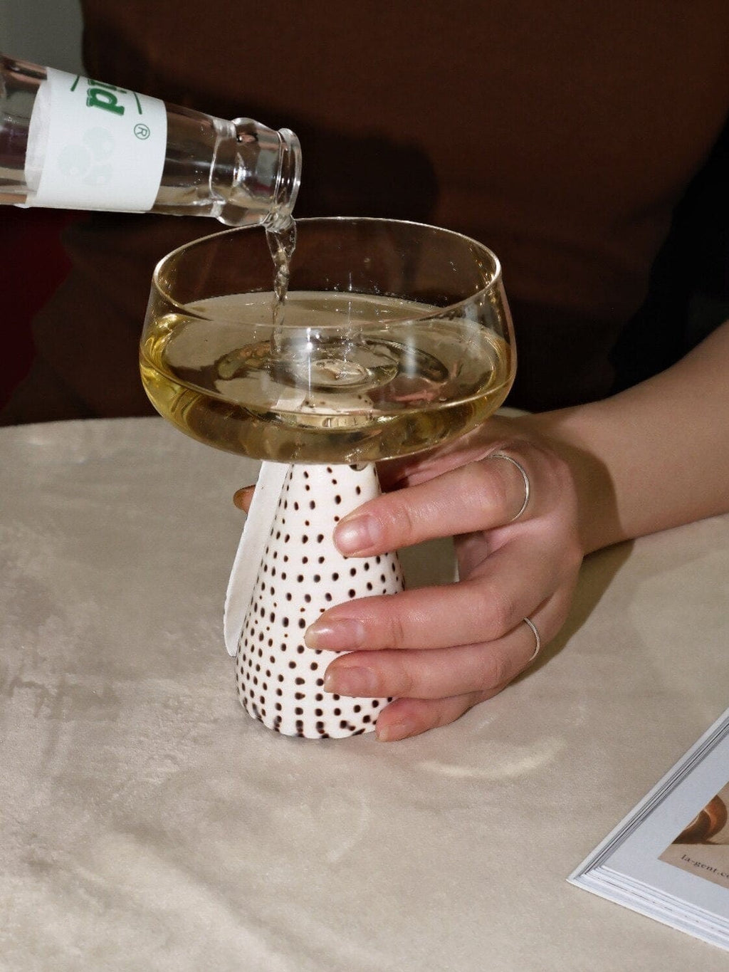 A white ceramic conch shell coupe glass with black polka dots held as golden liquid is poured from a bottle