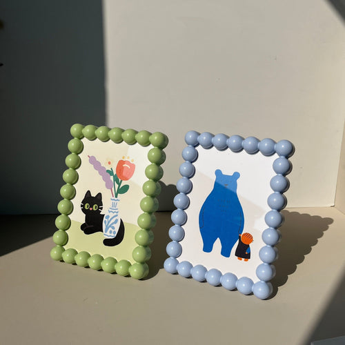 Two pastel-colored beaded retro picture frames with rounded bead borders featuring a black cat with flowers and a blue bear holding a figure