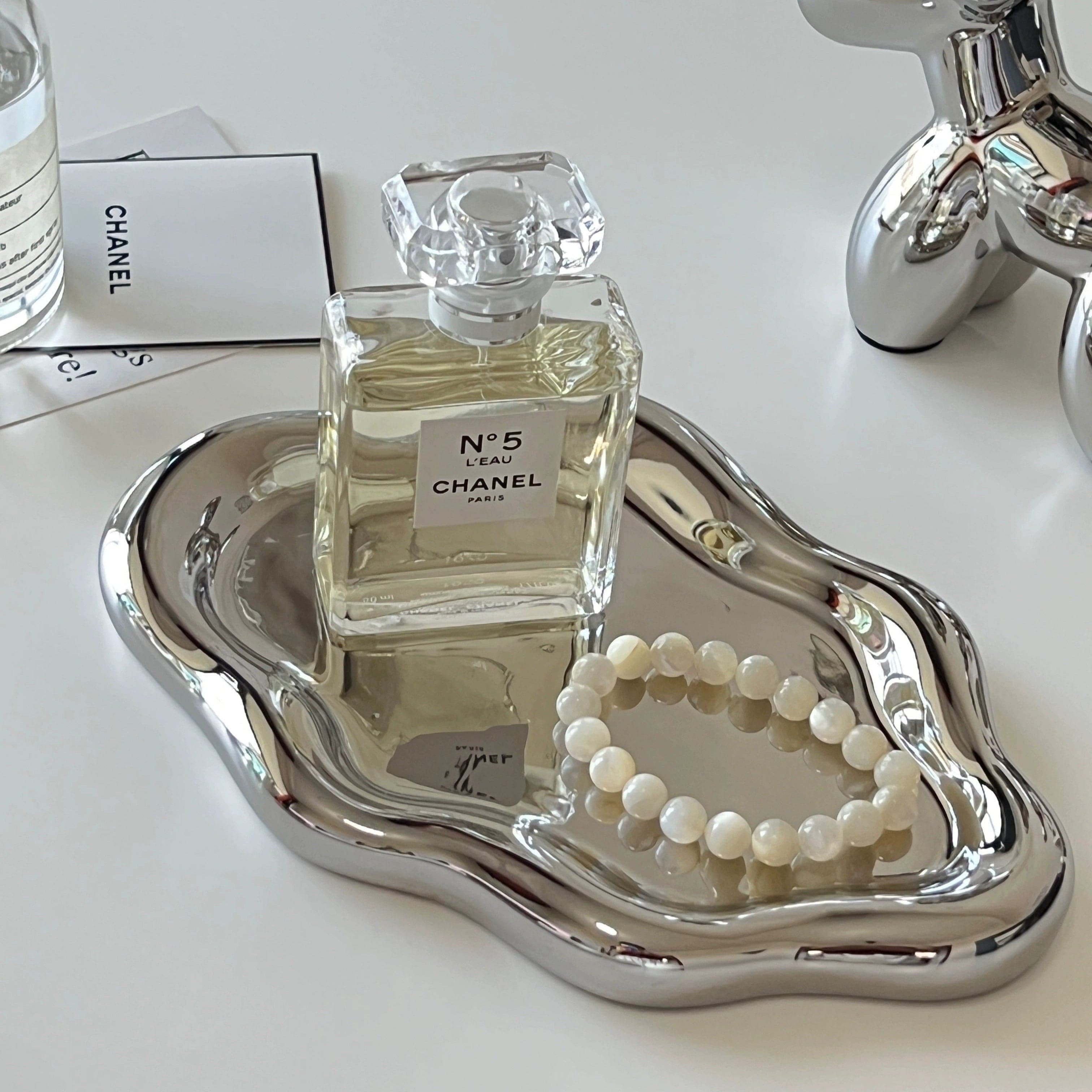 A sleek silver ceramic cloud tray with an organic shape holds a transparent glass bottle of Chanel No 5 perfume