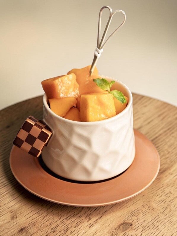 HOMELIVY white textured ceramic mug with a brown checkerboard cube handle, served with mango chunks on a saucer.