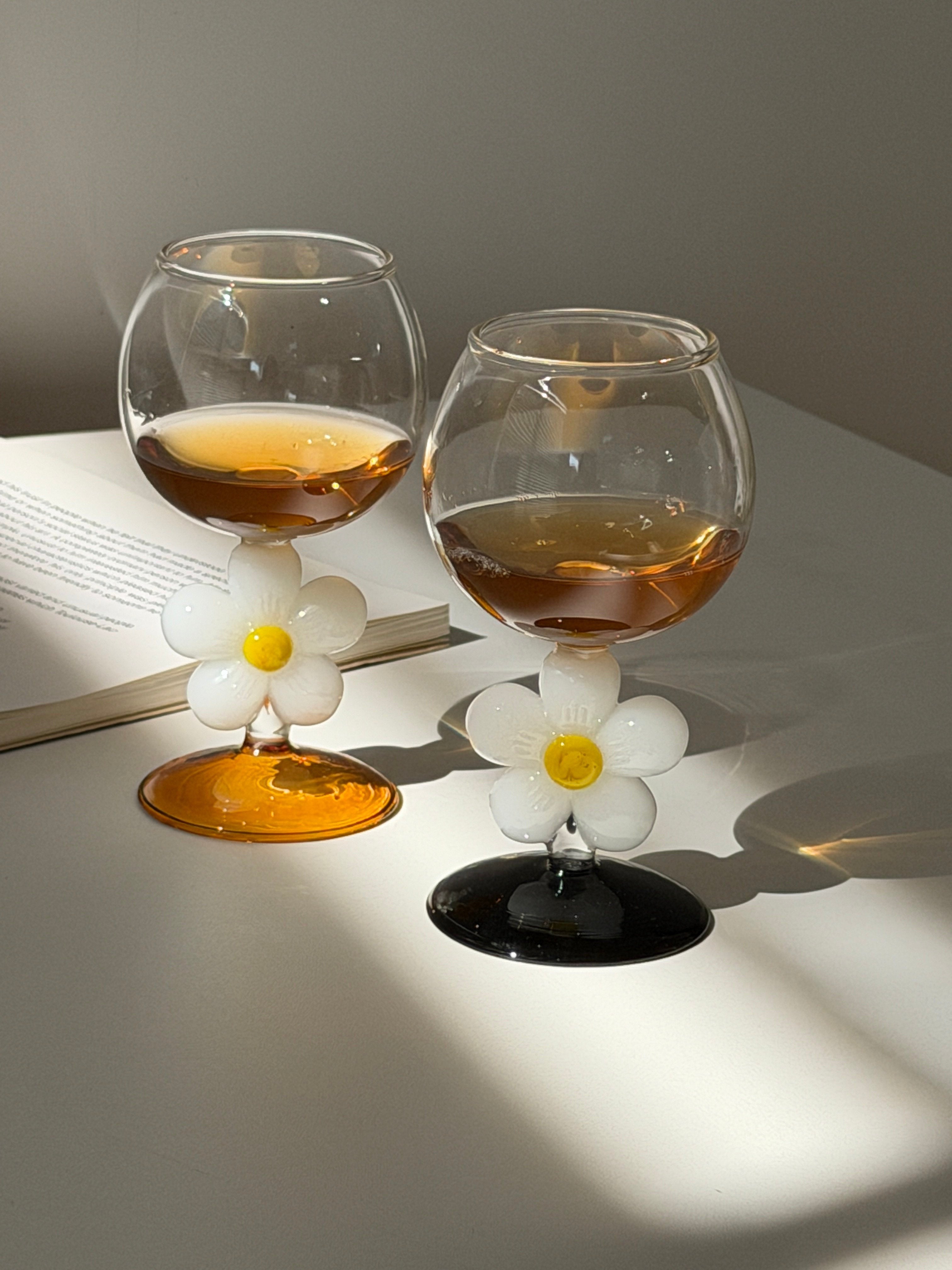 HOMELIVY daisy wine glasses with amber and black bases and white floral stems on a white table with a book.