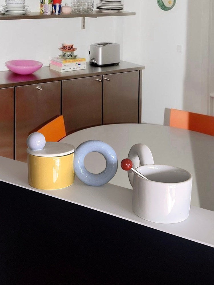 HOMELIVY ceramic donut handle mug in white with a red ball stirrer, displayed with a yellow lidded jar and blue ring.