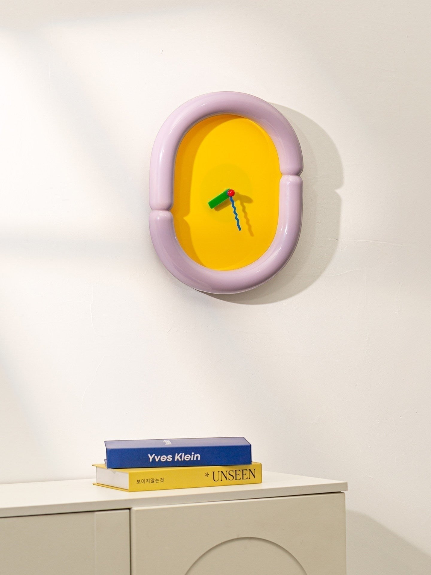 HOMELIVY oval wall clock with sculptural lilac frame and bright yellow face, featuring multicolored hands. A statement piece for eclectic decor.