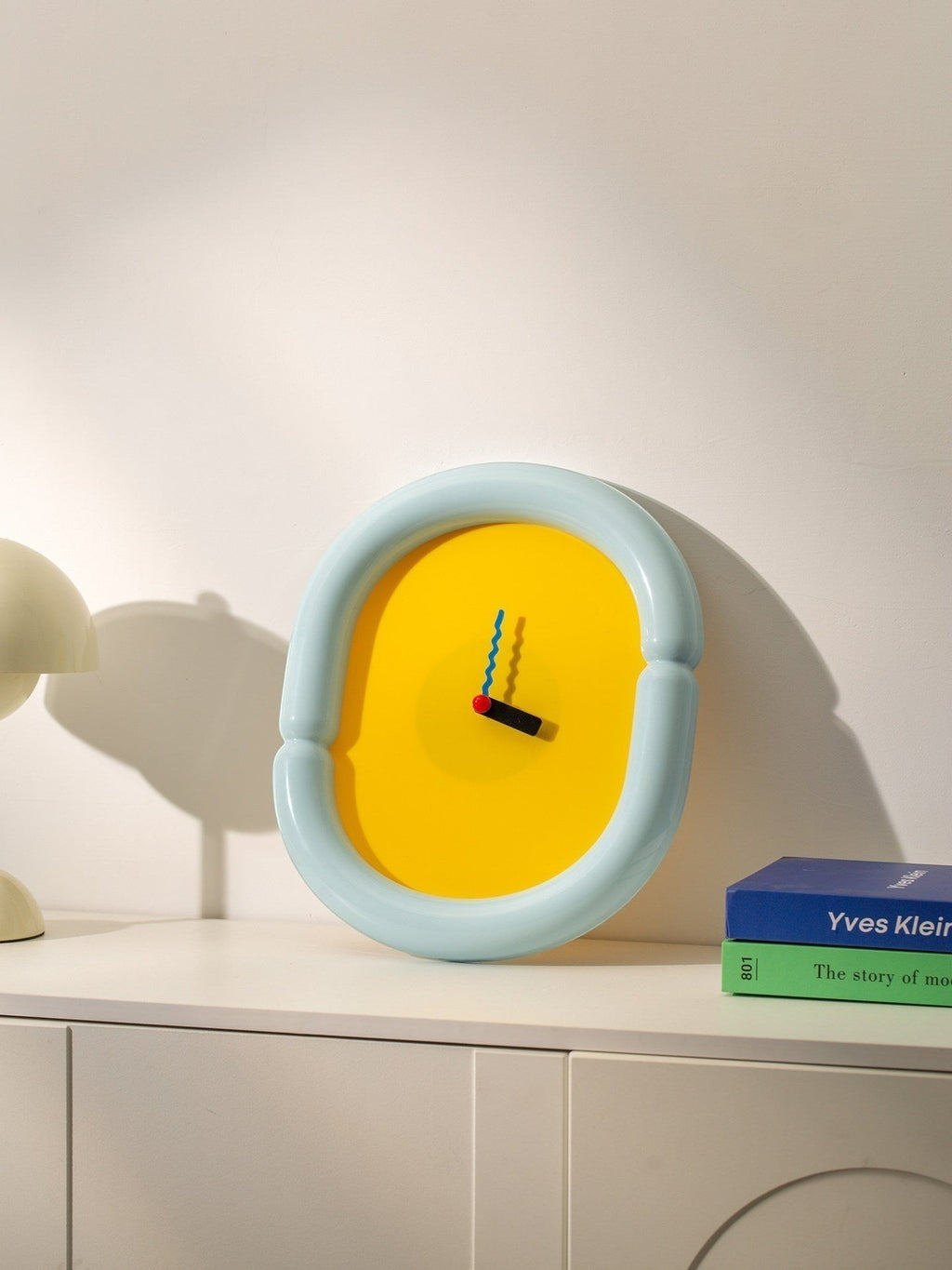 HOMELIVY sculptural wall clock with light blue frame and bold yellow face, featuring multicolored hands. A statement piece for eclectic decor.