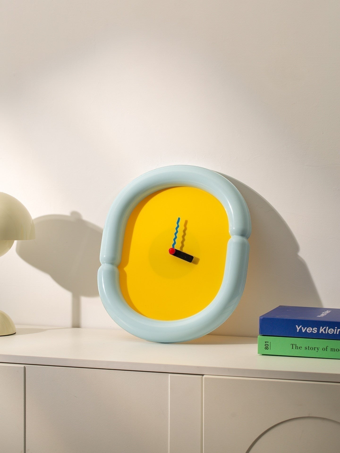 HOMELIVY sculptural wall clock with light blue frame and bold yellow face, featuring multicolored hands. A statement piece for eclectic decor.