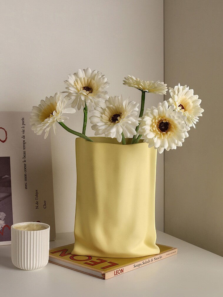 HOMELIVY yellow ceramic vase shaped like a paper bag, holding white daisies on a table with books and a candle.