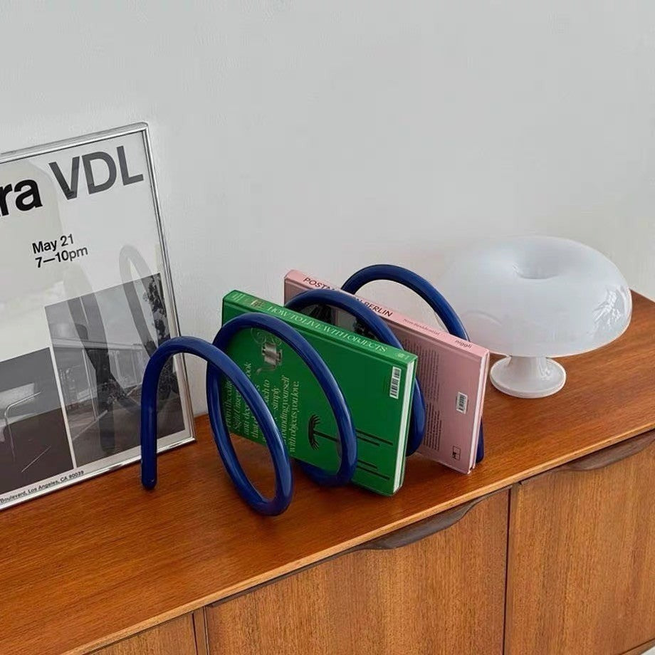 HOMELIVY blue spiral book holder displaying books on a wooden sideboard with a modern lamp and framed poster.