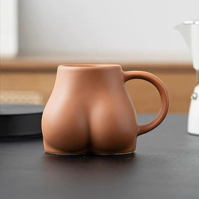 HOMELIVY Booty Mug, a cheeky butt-shaped ceramic coffee cup in a matte terracotta finish on a dark table.