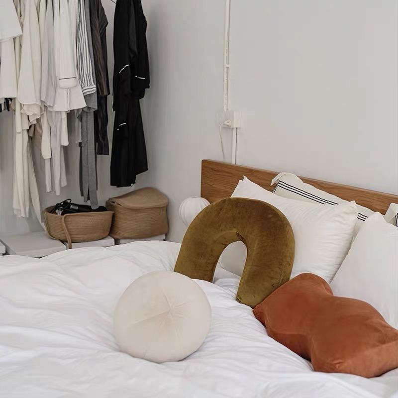 A plush olive green u-shaped neck pillow made of soft material resting on a bed with white and rust pillows